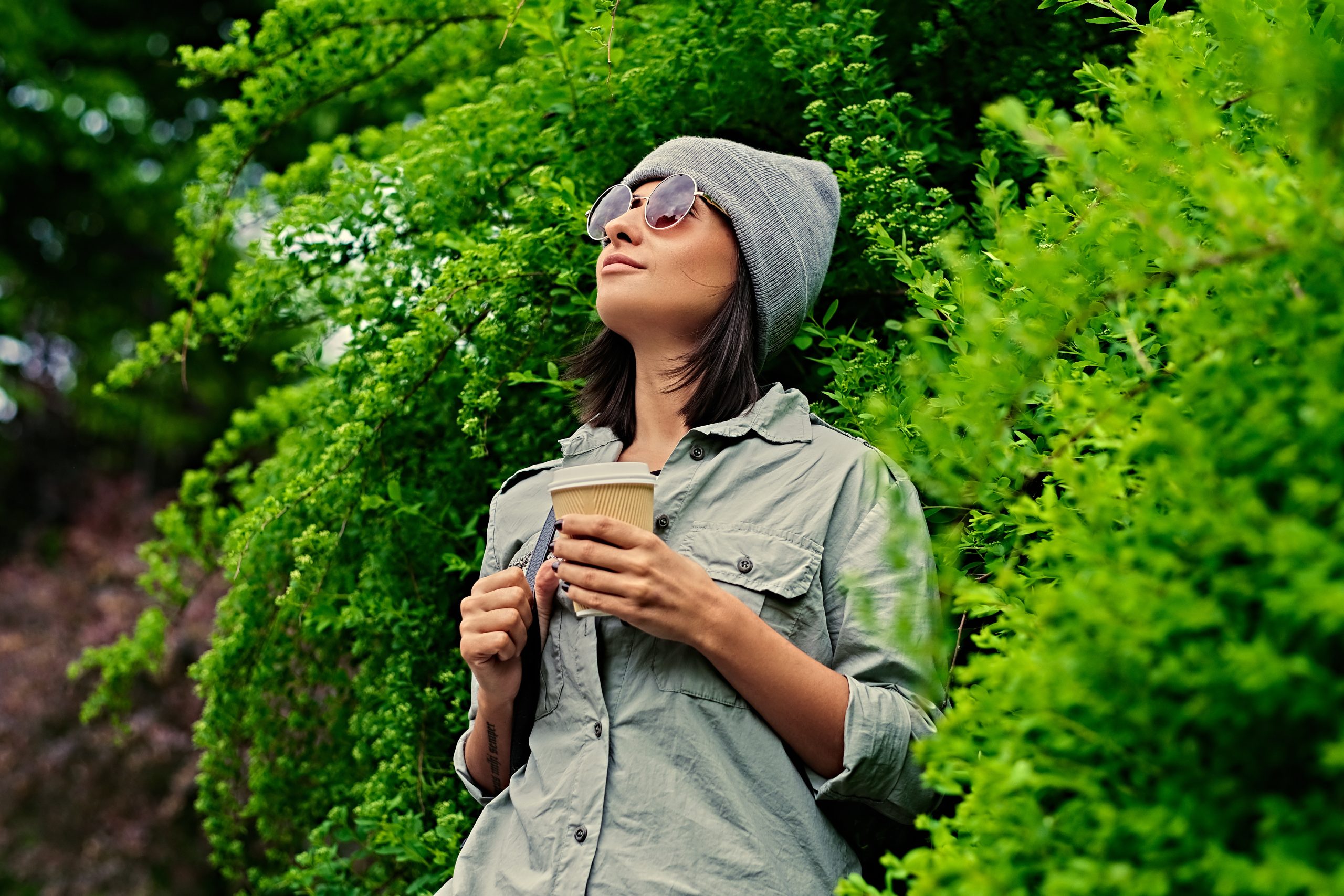 Portrait of young attractive female in sunglasses holds paper coffee cup in a green summer park.